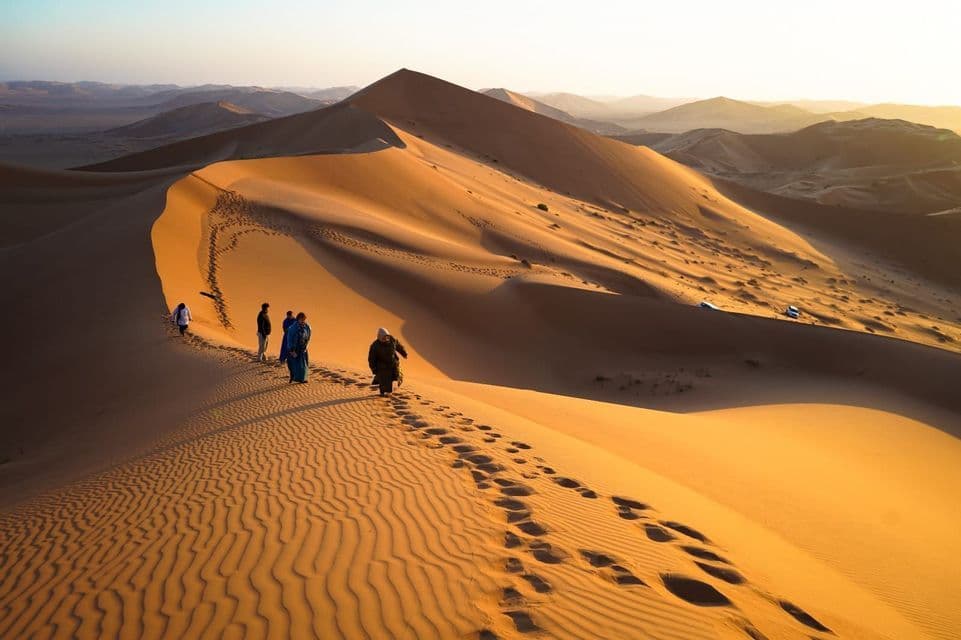 Un gruppo WeRoad cammina in fila lungo la cresta di una vasta duna di sabbia dorata, con montagne visibili in lontananza.