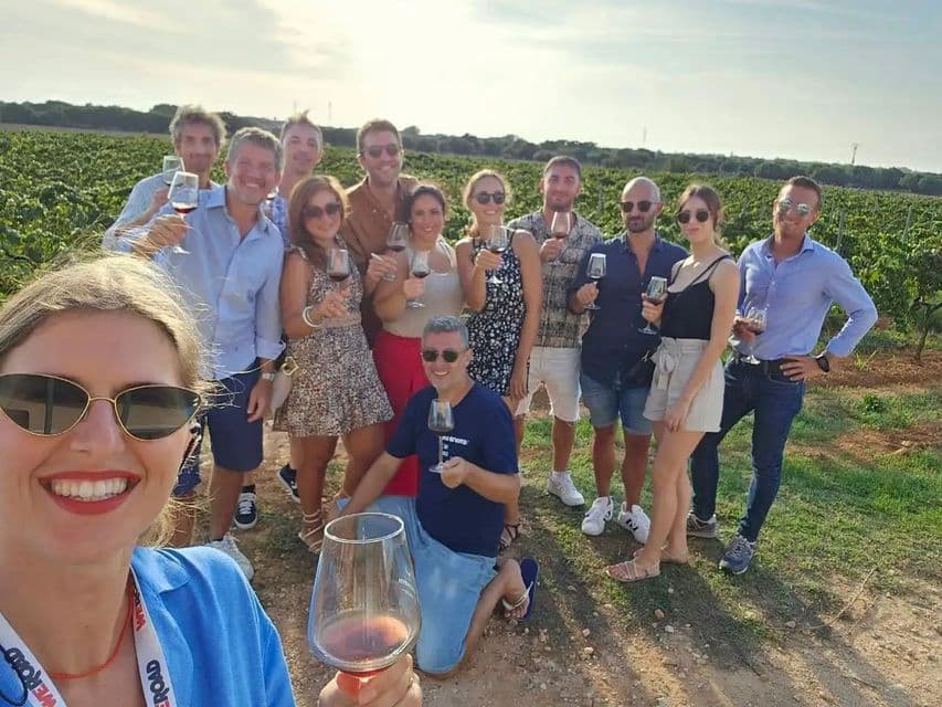 Un groupe WeRoad en voyage sourit pour une photo, verres de vin rouge à la main, dans un vignoble ensoleillé.