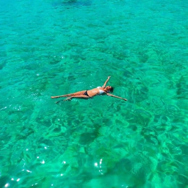 Une femme en bikini flottant sur le dos dans une eau claire et turquoise, vue d'en haut.