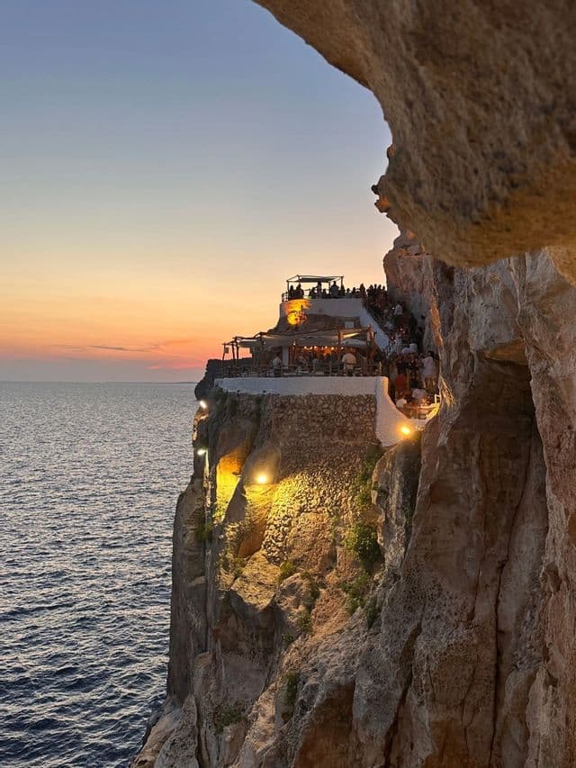 Un bar à plusieurs niveaux creusé dans une falaise rocheuse avec vue sur l'océan, visible depuis l'entrée d'une grotte au coucher du soleil.