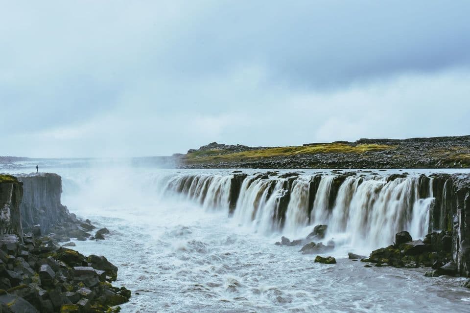 Una persona solitaria se alza sobre un acantilado rocoso, empequeñecida por una ancha y potente cascada bajo un cielo nublado.