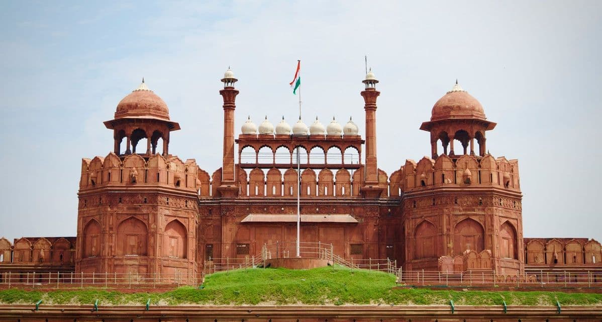 La bandiera dell'India sventola sopra i bastioni e le cupole di arenaria rossa di una grande fortezza simmetrica sotto un cielo coperto.