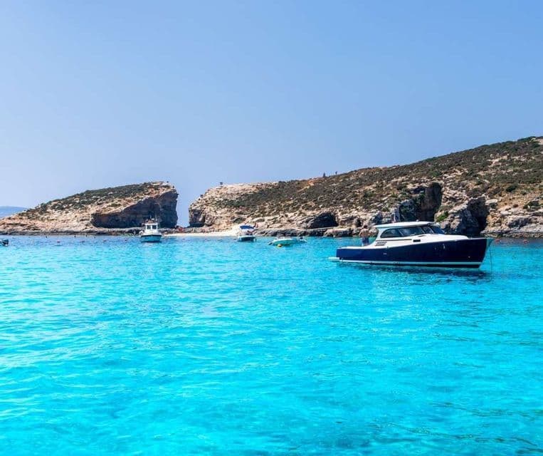 Several boats float on clear turquoise water near a rocky coastline under a bright blue sky.