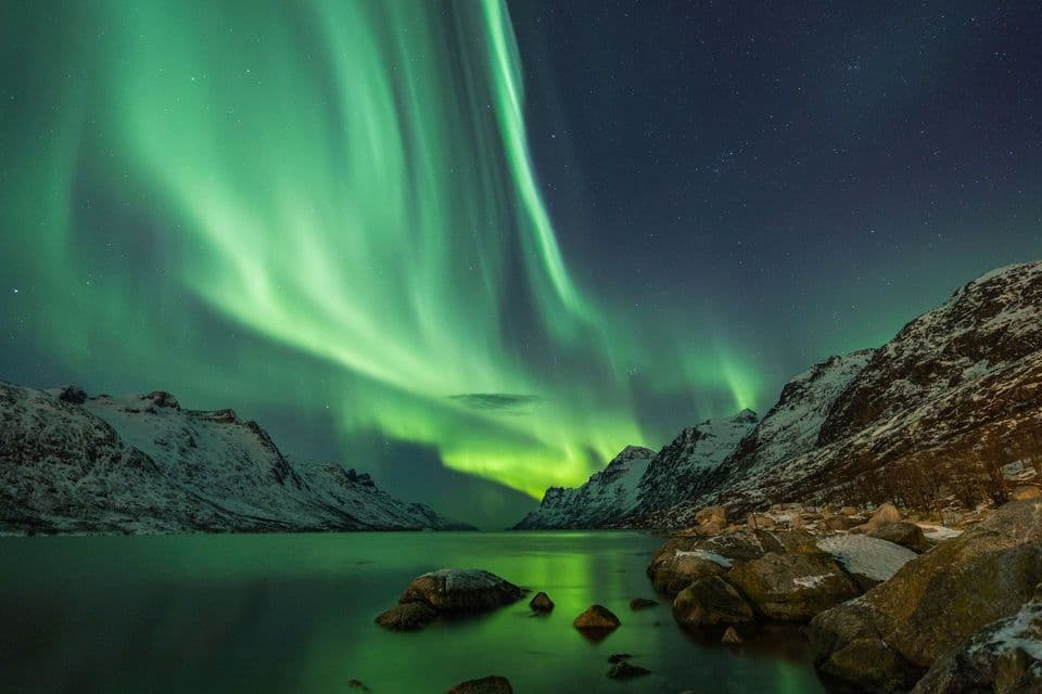 L'aurora boreale verde illumina un cielo notturno stellato sopra montagne innevate e un lago calmo e riflettente.