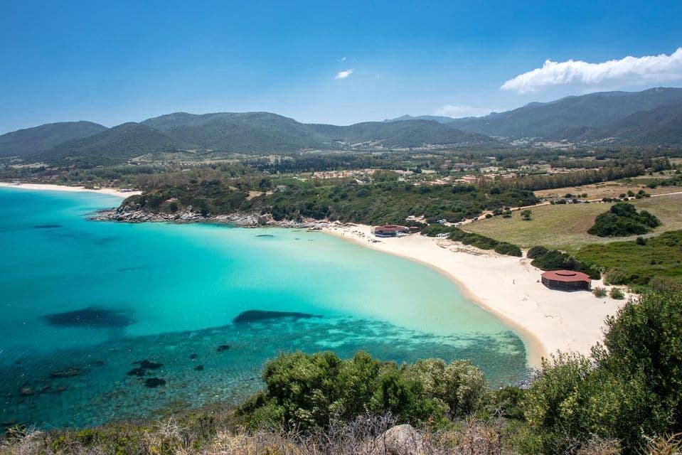 Vue en plongée d'une plage de sable serpentant le long d'un littoral, avec une eau turquoise claire, bordée par des collines verdoyantes sous un ciel bleu.