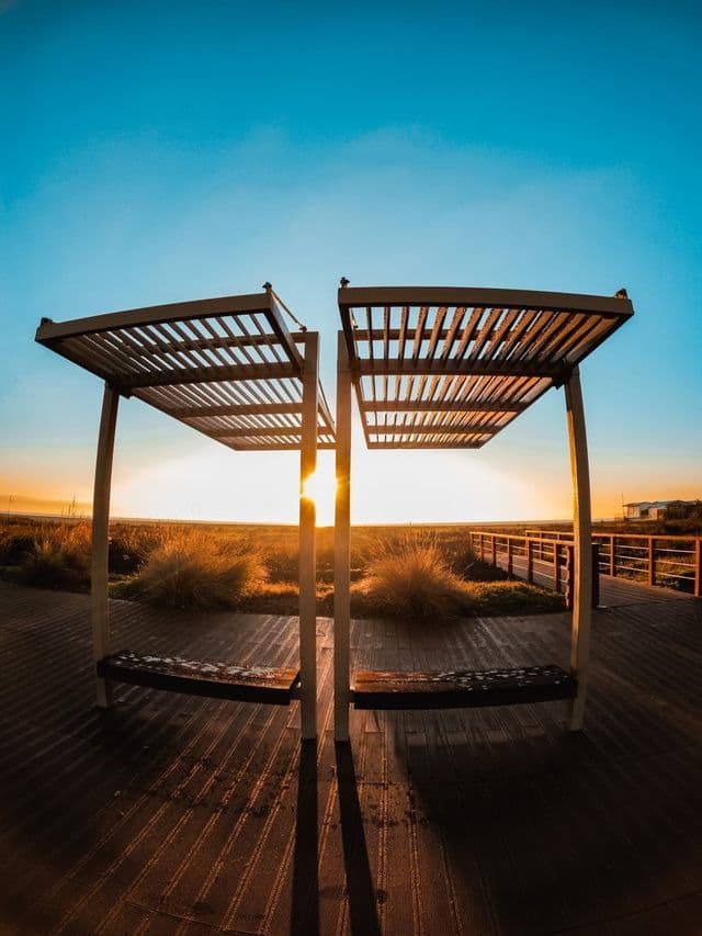 Dos bancos modernos con pérgolas de lamas en un paseo de madera, frente a un paisaje herboso durante un atardecer dorado bajo un cielo despejado.