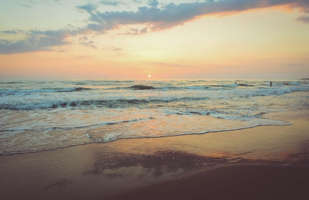 El sol se pone sobre el océano mientras las olas rompen en una playa de arena, con siluetas de personas surfeando en la distancia.