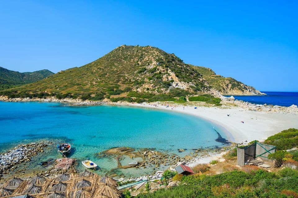 Una vista aérea de una cala recóndita con una playa de arena blanca y agua turquesa, enclavada al pie de una gran colina verde.