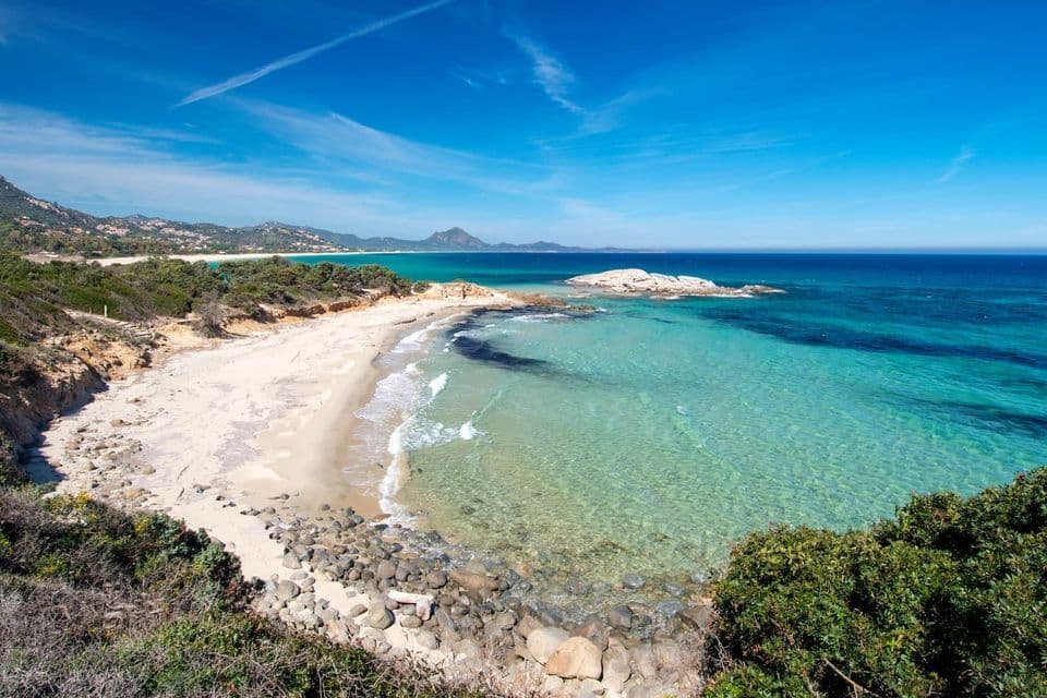 Ein malerischer Blick auf einen abgelegenen weißen Sandstrand mit klarem türkisfarbenem Wasser, umrahmt von grüner Vegetation unter einem weiten blauen Himmel.
