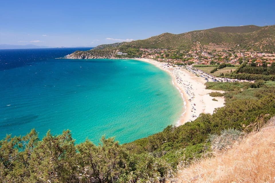 Vista aérea de una playa arenosa y concurrida que bordea una bahía de aguas turquesas, con un pueblo costero y colinas verdes al fondo.