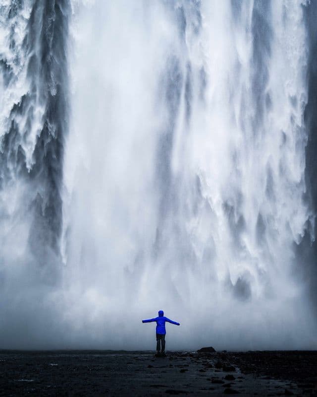 A person in a blue jacket with arms outstretched stands at the base of a massive waterfall.