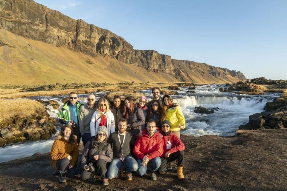 A WeRoad group trip poses for a photo in front of a rushing river at the base of a rocky, sunlit cliffside.