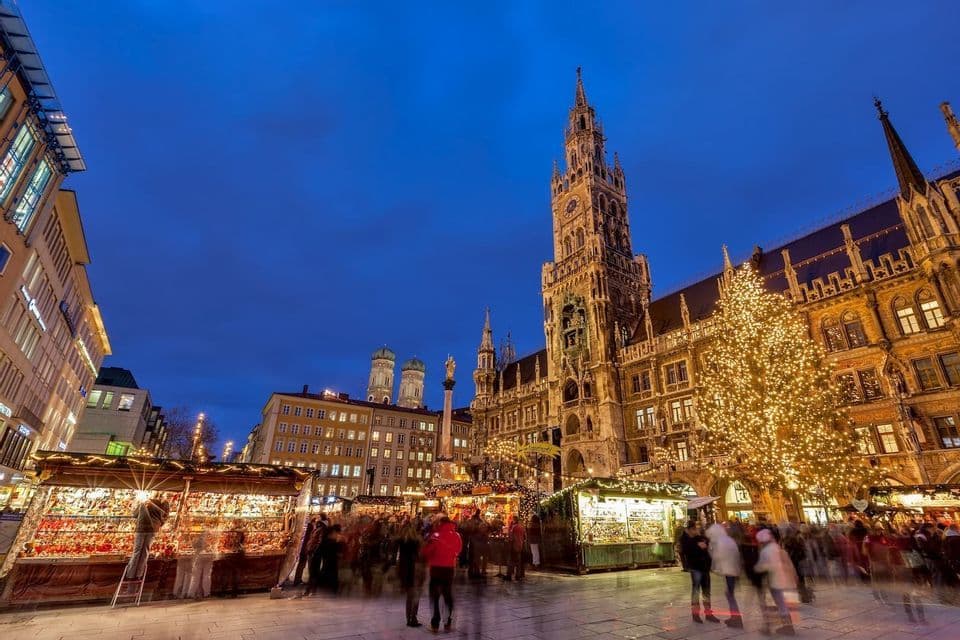 Ein geschäftiger Weihnachtsmarkt auf einem historischen Stadtplatz bei Dämmerung, mit einem beleuchteten Baum und einem verzierten Uhrturm vor einem tiefblauen Himmel.