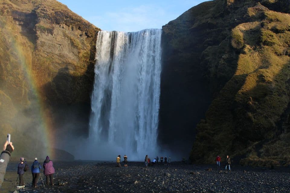 A WeRoad group trip stands on a rocky shore looking at a massive waterfall, with a rainbow visible in the mist.