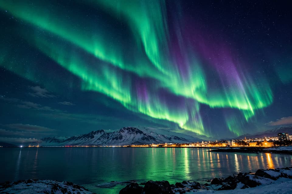 Aurora boreale verde e viola illumina il cielo stellato sopra una città costiera e montagne innevate, con riflessi nell'acqua.