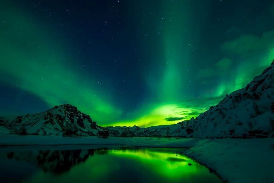 L'aurora boreale verde brilla in un cielo notturno stellato sopra montagne innevate, riflettendosi in un lago calmo.