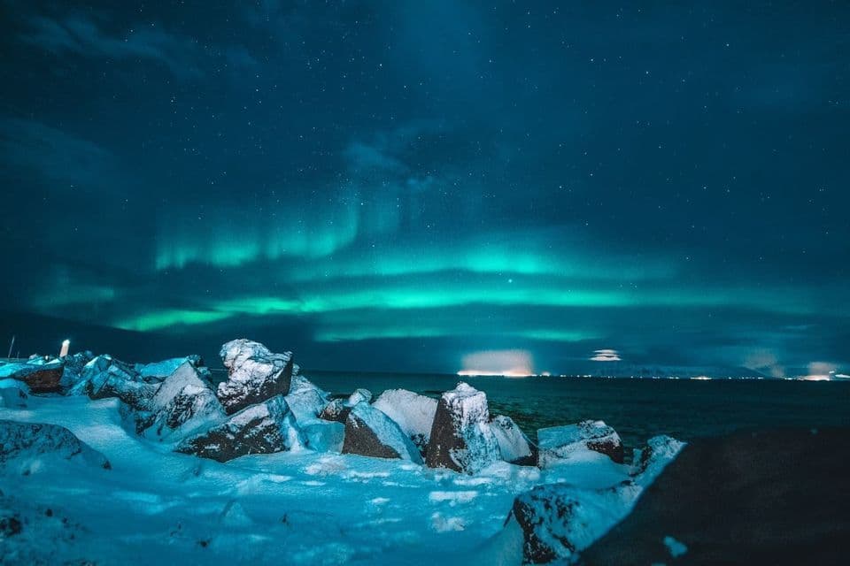 L'aurora boreale risplende nel cielo notturno stellato, sopra una riva di rocce innevate e acqua.