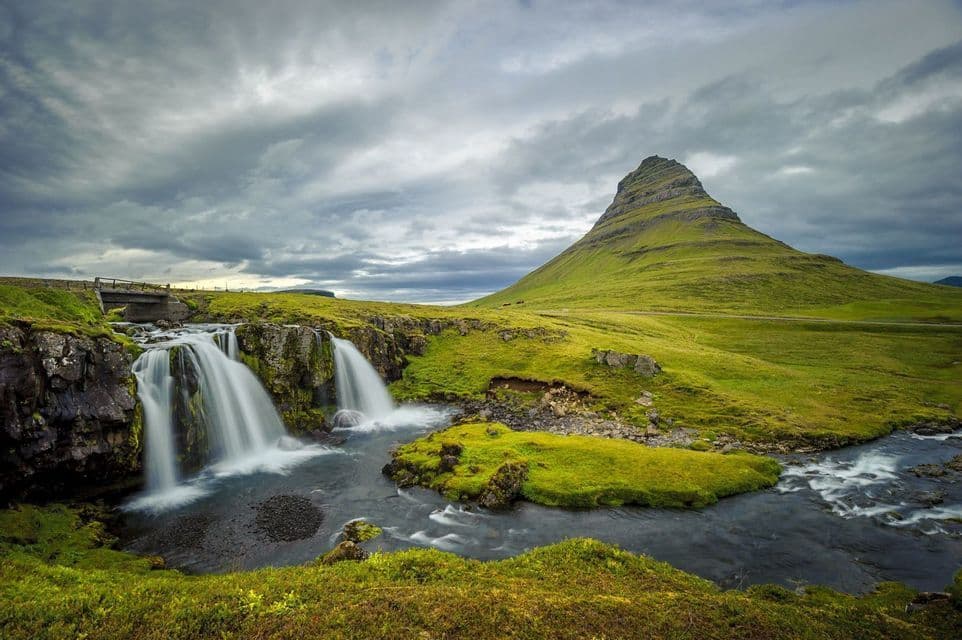 A waterfall cascades down rocks into a river, with a large, green conical mountain in the background under a cloudy sky.