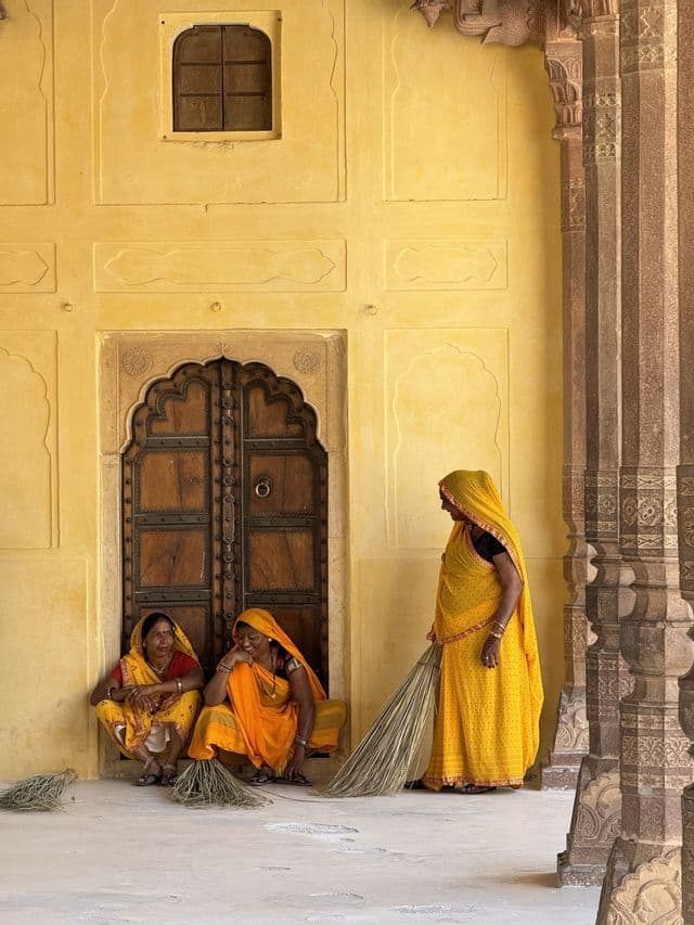 Tre donne in sari gialli si concedono una pausa dalla pulizia. Due sono sedute accanto a una porta di legno, e una è in piedi con una scopa contro un muro giallo.