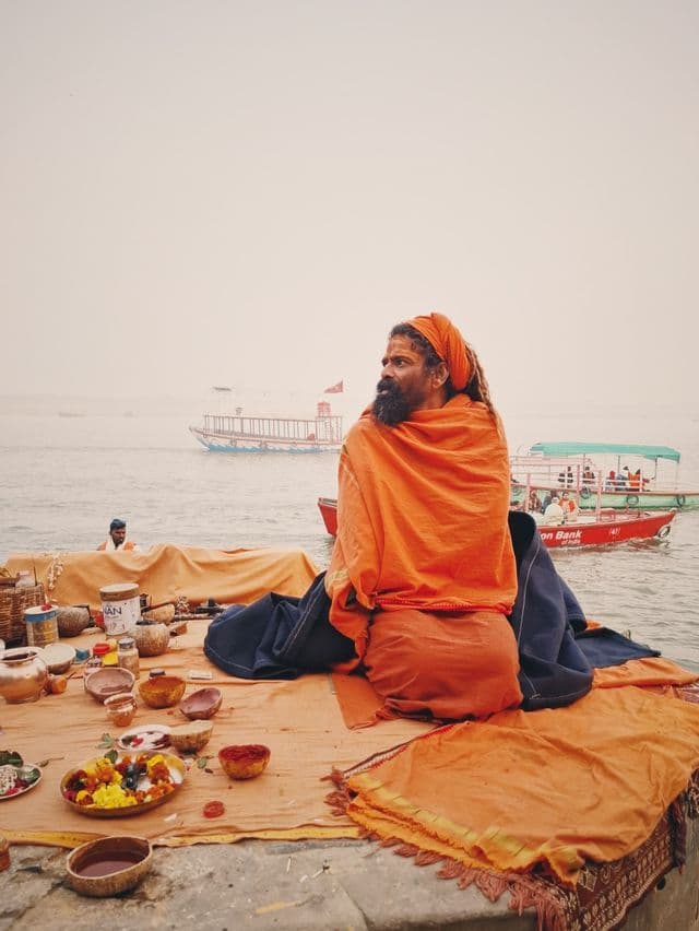 Un hombre barbudo con rastas, vestido con túnicas naranjas, sentado en una plataforma junto a un río brumoso, con ofrendas rituales dispuestas ante él.