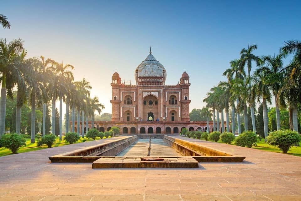 Un mausoleo in arenaria rossa con una grande cupola alla fine di una lunga vasca riflettente, affiancato da palme sotto un cielo sereno.