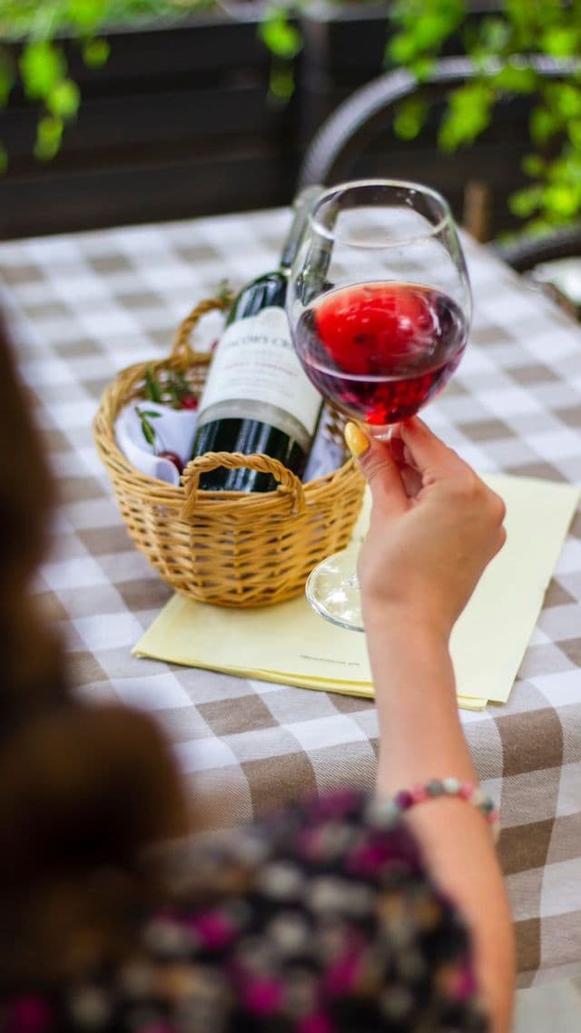 A person's hand holds a glass of red wine over a checkered tablecloth, with a bottle of wine in a wicker basket.