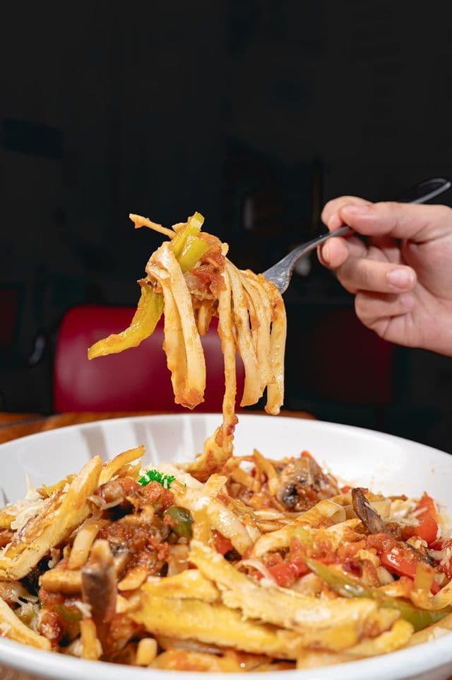 A hand lifts a forkful of fettuccine pasta with vegetables and sauce from a white bowl in a restaurant setting.