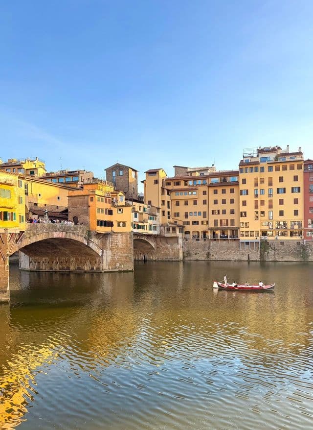 A WeRoad group trip in a red boat on a river beside a historic stone bridge and yellow buildings under a clear sky.