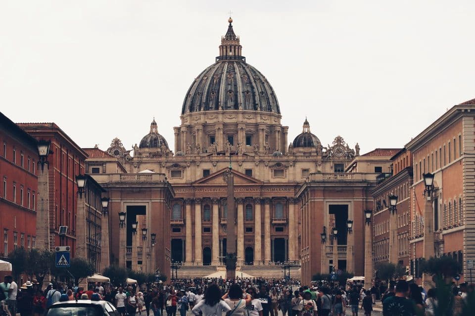 A large crowd of people on the street in front of the grand facade and large dome of St. Peter's Basilica under an overcast sky.