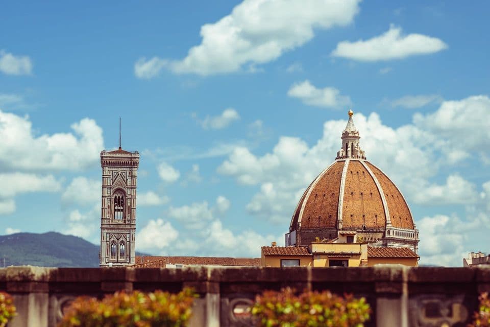 A large terracotta dome and an ornate bell tower stand against a blue sky with white clouds, viewed over a stone wall.