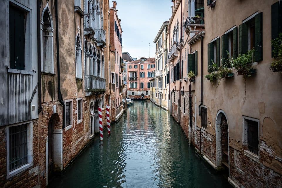 A narrow canal flows between tall, historic buildings with arched windows and balconies, reflecting the overcast sky.