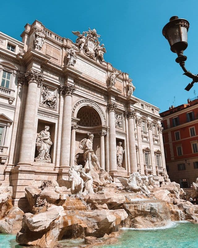 A grand, ornate fountain featuring classical sculptures with water flowing into a turquoise basin under a bright blue sky.