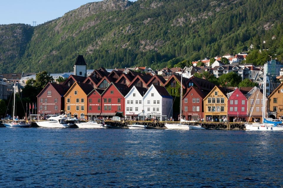 Colorful wooden buildings line a waterfront harbor with moored boats beneath a forested mountain slope.