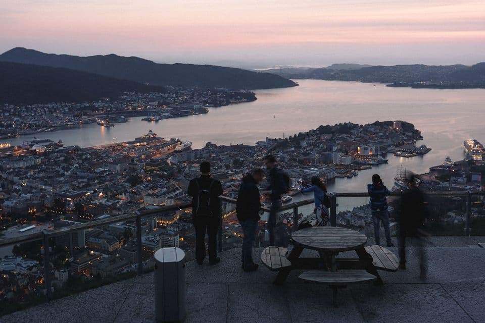 People standing at an elevated viewpoint overlooking a coastal harbor town surrounded by hills during twilight.