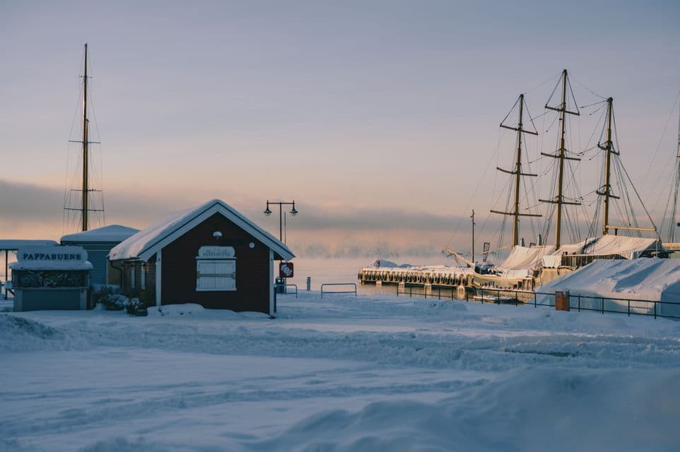 Snow-covered wooden cabin and docked sailing ships at a tranquil harbor under a pale winter sky.