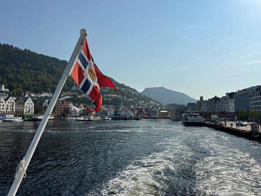 A flag flutters from the stern of a boat sailing away from a waterfront harbor town surrounded by forested mountains.