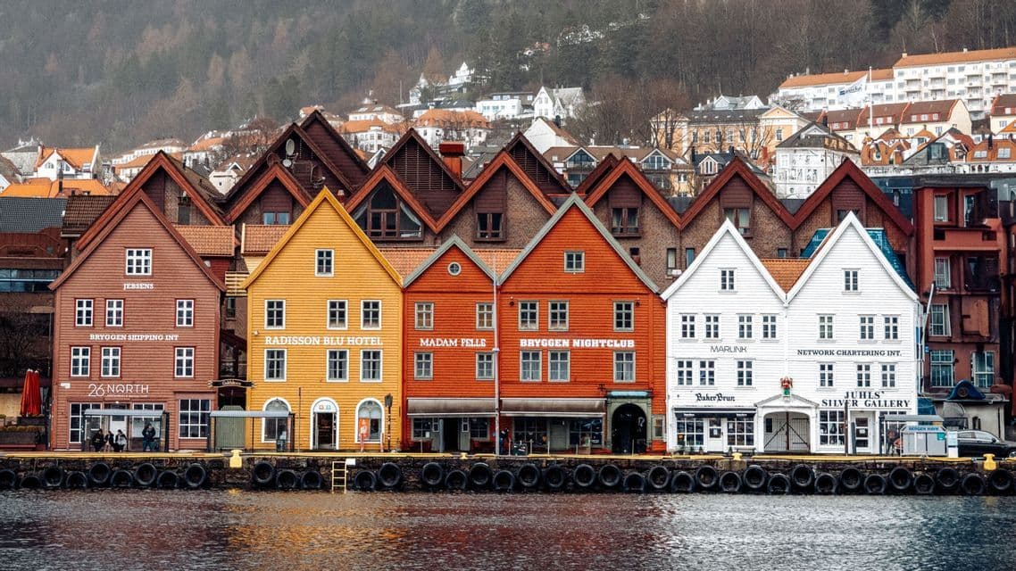 Colorful wooden gable-roofed buildings line a waterfront harbor with hillside houses in the background.