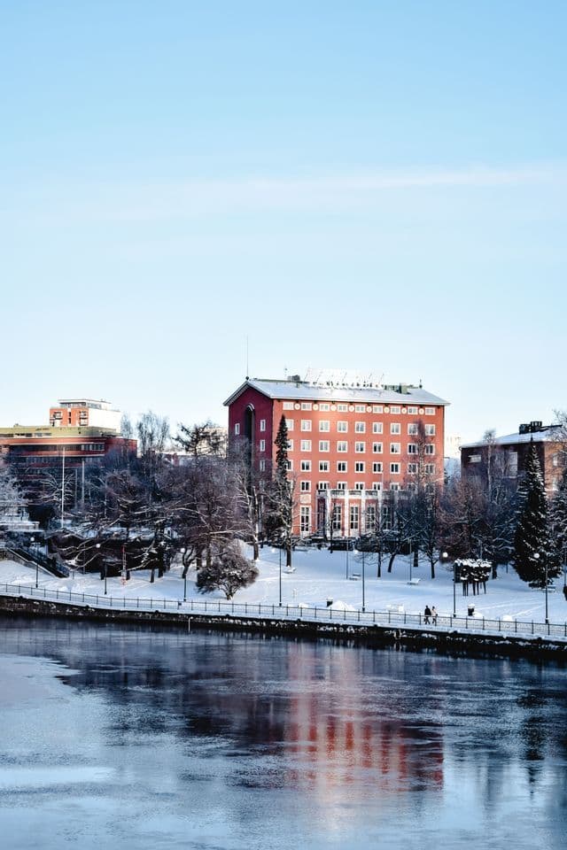 A red multi-story building stands along a snow-covered riverbank, casting reflections in the cold water.