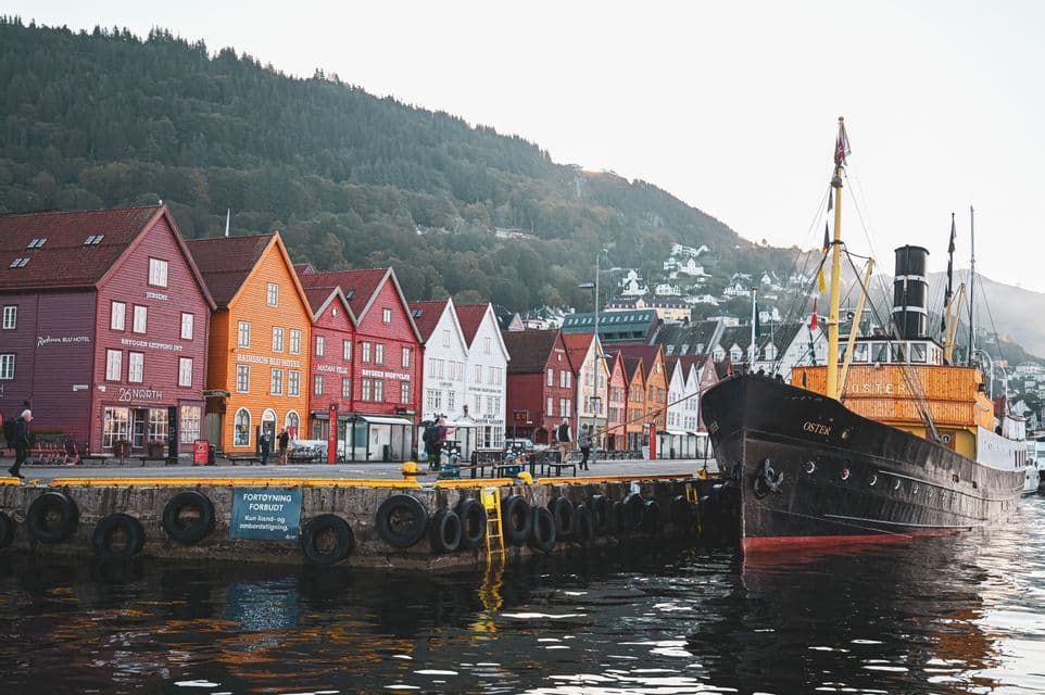 A vintage ship docked at a pier beside colorful wooden waterfront buildings with forested hills in the background.