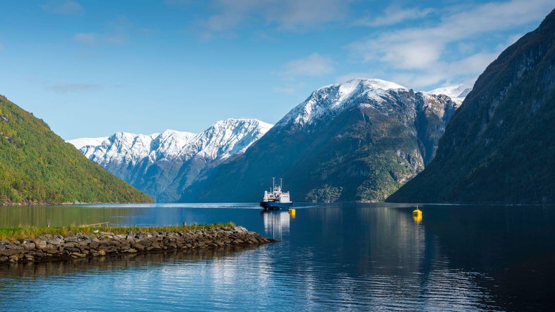 A ferry navigates calm blue waters surrounded by steep, snow-capped mountains under a clear sky.