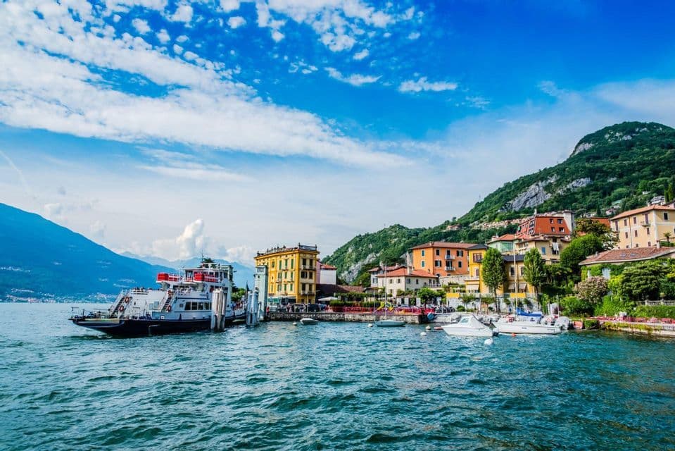 A ferry docked at a pier in a colorful lakeside town at the base of a green mountain under a blue, cloudy sky.