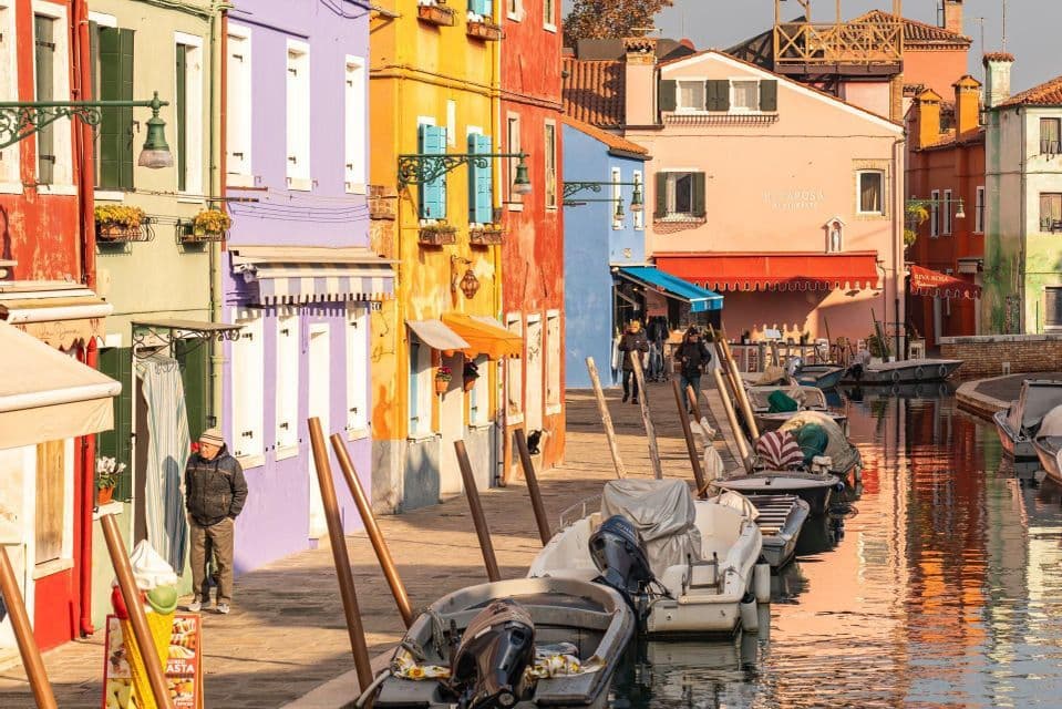 Colorful houses line a sunlit canal where boats are moored and people walk along the pavement.