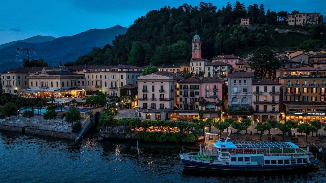 A colorful lakeside town is illuminated at dusk, with a boat on the water and a forested mountain rising in the background.