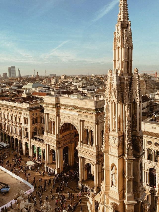 An ornate gothic spire overlooks a grand arched building and a bustling city square from a high-angle perspective under a blue sky.