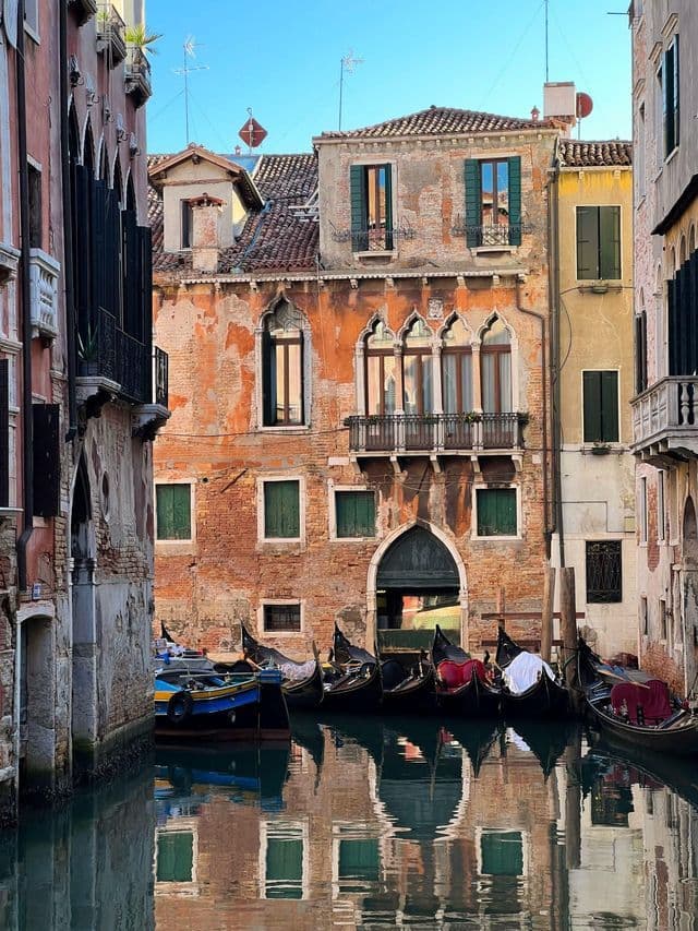 Gondolas moored in a narrow canal, with reflections of old brick buildings on the calm water under a clear blue sky.