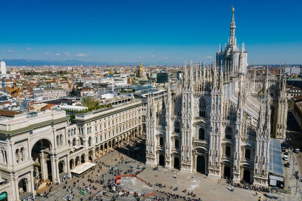 An aerial view of a large, ornate Gothic cathedral and an adjacent public square bustling with people under a clear blue sky.