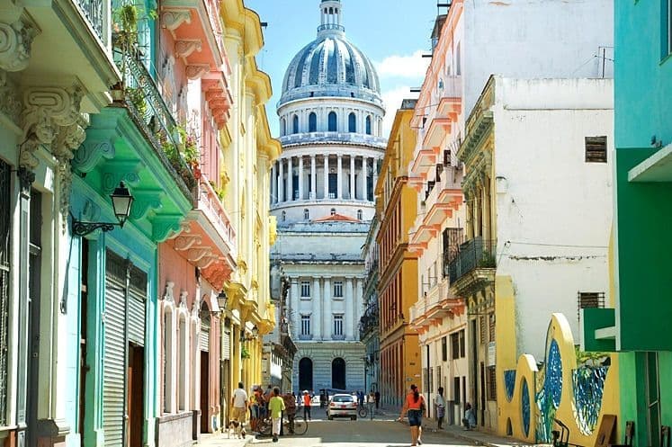 Una strada cittadina fiancheggiata da edifici colorati conduce verso un grande edificio capitolino a cupola in stile classico sotto un cielo limpido.
