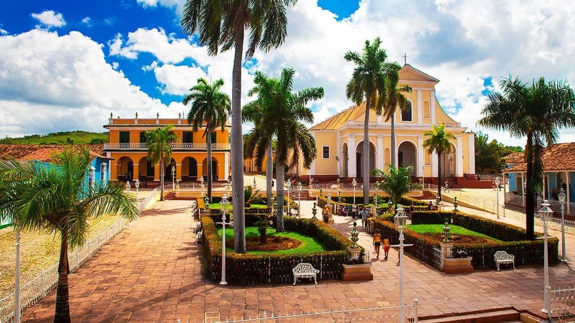 Una piazza cittadina soleggiata caratterizzata da edifici coloniali gialli, alte palme e un giardino centrale sotto un cielo azzurro brillante con nuvole.