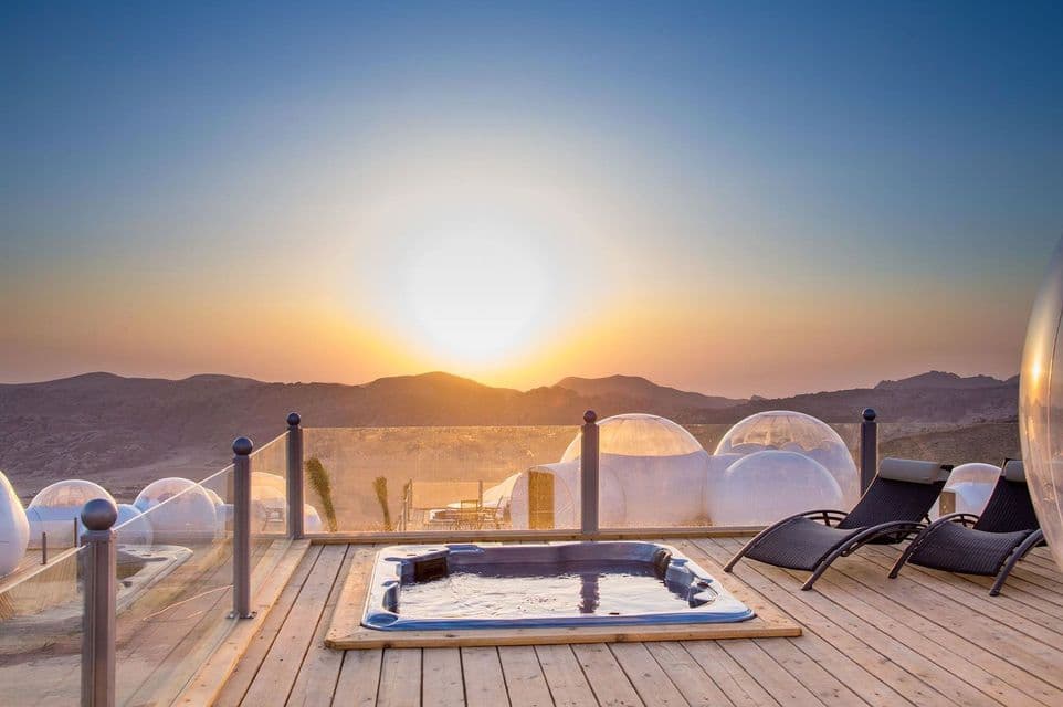 Tiendas domo burbuja en una terraza de madera con tumbonas y un jacuzzi, con vistas a un paisaje montañoso desértico.