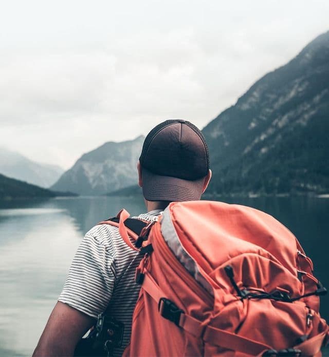 Una persona con mochila naranja y gorra contempla un lago sereno rodeado de montañas brumosas.
